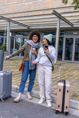Female friends laughing and checking phone outside terminal with luggage and coffee