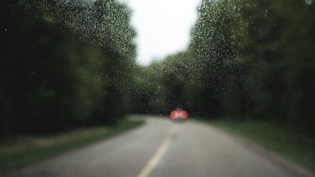 Raindrops on car window offering a dynamic perspective of a rural road and lush green trees and countryside in the background. The image's artistic, blurred effect is suitable for visual themes of travel, weather conditions, long drives, or nature's beauty. It can be used in blogs, travel websites, environmental content, and scenic backgrounds conveying a sense of calm and reflection.