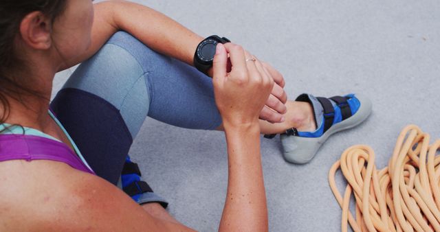 Woman checking smartwatch while sitting on ground, relaxing after rock climbing session, climbing gear beside her. Ideal for content on fitness, wearable technology, outdoor sports, healthy lifestyle.