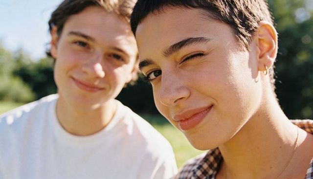 Winking short-haired teen wearing gold hoop leaning forward with smiling friend in sunlit park close