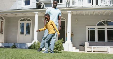 African american father guiding son on suburban front lawn near white porch and seating