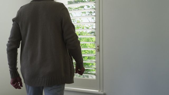 Elderly man stands in bedroom facing window, with sunlight streaming and greenery visible outside. Man wears casual sweater and jeans, creating a tranquil and introspective scene. Suitable for themes of solitude, retirement, introspection, peaceful living, and daily home routines.