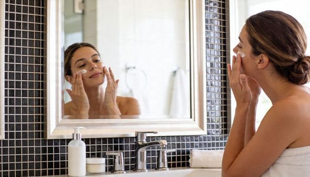 Towel-wrapped woman applying moisturizer while looking in mirror at modern bathroom vanity