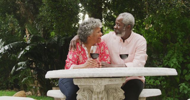 Senior biracial couple enjoys a moment outdoors, with copy space. They share a romantic wine toast in a serene garden setting.