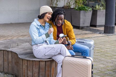 Young travelers sharing phone and coffee while waiting with luggage in urban plaza
