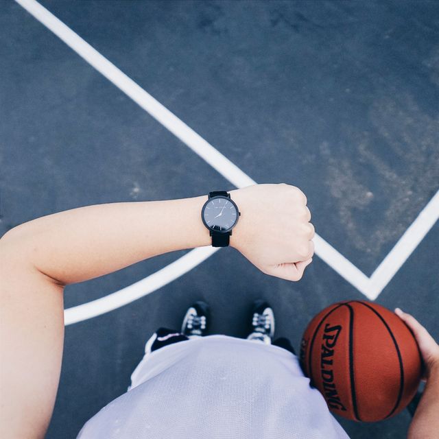 Photo shows a close-up of a person holding a basketball on a sports court, while looking at their wristwatch. Can be used for themes related to sports, time management, athletic preparation and focus.