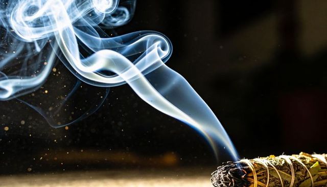Smoldering sage bundle releasing swirling blue smoke with ember sparks and warm glow