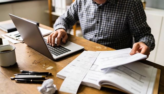 Man sorting receipts while typing on laptop at wooden desk for bookkeeping and tax prep
