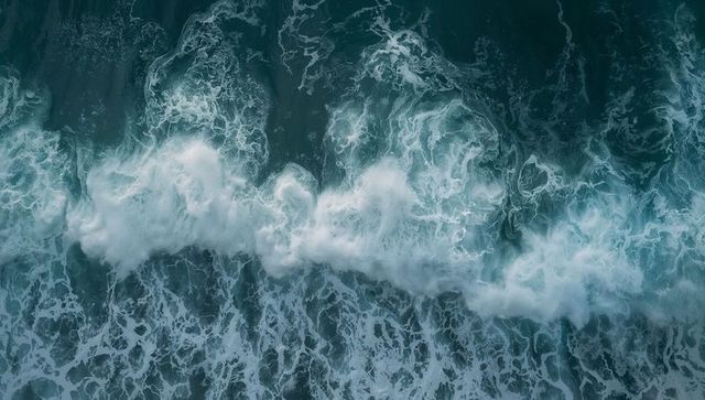 Aerial breaking ocean wave churning dense white foam revealing swirling teal currents
