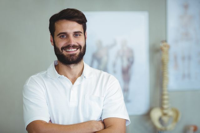 Male physiotherapist standing with arms crossed, smiling confidently in a clinic. Ideal for use in healthcare, medical, and wellness-related content, showcasing professional physiotherapy services and patient care.
