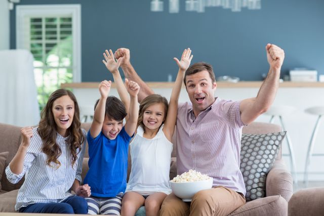 Family of four sitting on couch in living room, cheering and smiling while watching TV. Parents and children are enjoying time together, holding a bowl of popcorn. Perfect for advertisements, family-oriented content, and articles about family bonding, home entertainment, and leisure activities.