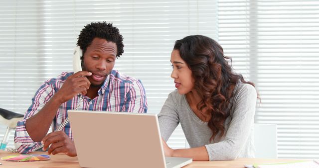 Two coworkers collaborating at an office desk. They are actively discussing something important, illustrating teamwork and communication. The man is on the phone while the woman is focused on the laptop screen. This scene can be used in contexts related to business meetings, office communication, teamwork, project planning, and corporate training materials.