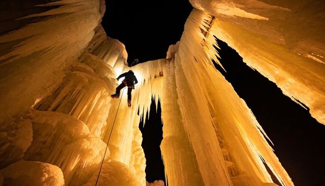 Night ice climbing in amber frozen canyon with climber lowering on rope under stalactites