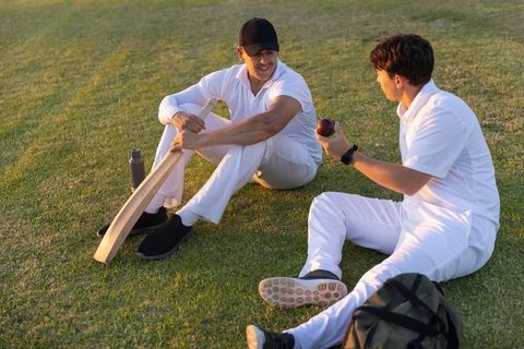 Cricket teammates sitting on grass sharing bat and ball during golden hour