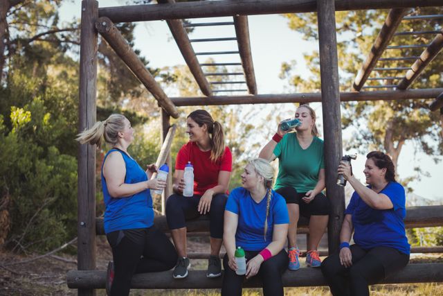 Group of women taking a break and hydrating during an outdoor boot camp on a sunny day. They are sitting on a wooden structure, holding water bottles, and engaging in conversation. Ideal for use in fitness, health, and wellness promotions, as well as advertisements for outdoor activities and group exercise programs.