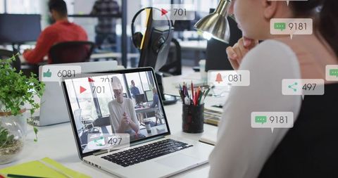 Young professional watching live video call on laptop in open office with engagement overlays
