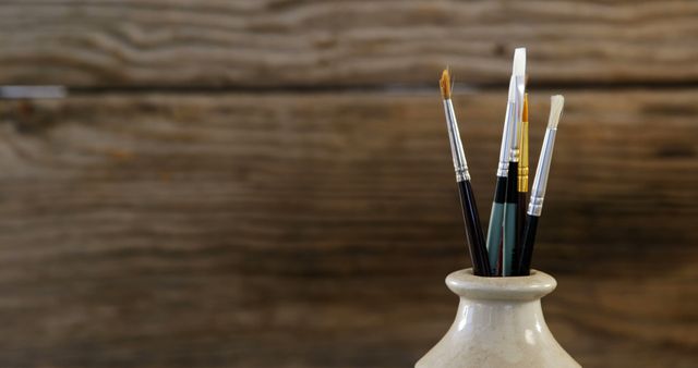 Artist brushes neatly arranged in a ceramic vase on a rustic wooden background. Ideal for illustrating creative workspaces, art studios, or showcasing artistic tools in vintage settings. Useful for blogs or websites about painting, art and crafts, or decorating ideas with an artistic touch.
