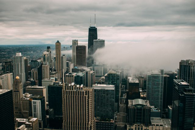Aerial view of Chicago's iconic skyline featuring a mix of modern and historic skyscrapers on a cloudy day. Thick clouds shroud parts of the buildings, adding a dramatic feel to the scene. Ideal for use in articles, travel guides, and promotional materials about Chicago, urban landscapes, architecture, and city life.
