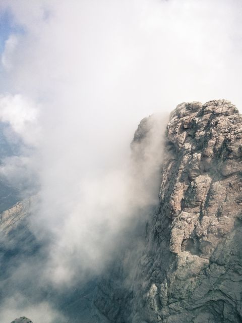 Misty mountain peak emerging majestically from the clouds, with rugged, rocky cliffs partially obscured by dense fog. Ideal for use in nature or adventure projects, travel blogs, posters, presentations, or backgrounds.
