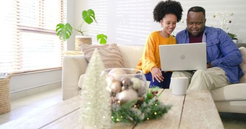 Couple sharing laptop on cozy sunlit sofa with warm holiday decor on wooden coffee table