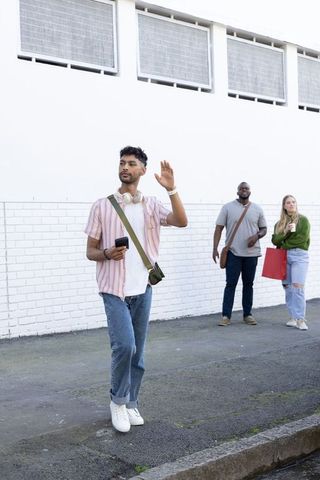 Young man waving while holding smartphone and wearing headphones, walking with friends