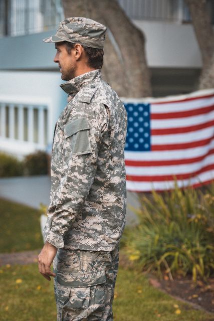 Soldier in camouflage uniform standing outdoors next to United States flag. Ideal for use in content related to military service, patriotism, homecoming celebrations, veterans, and national pride.