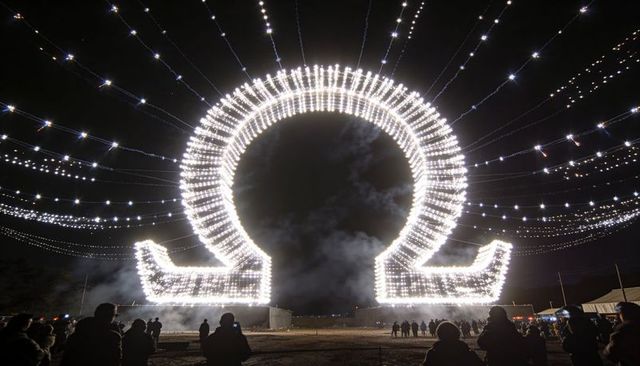 Omega light sculpture glowing over night festival crowd with radiant string lights