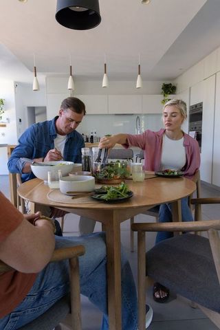 Group serving fresh salad around round wooden dining table in bright modern kitchen