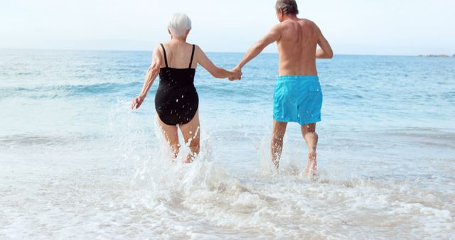 Senior couple enjoying beach vacation together, running into the ocean while holding hands. Useful for depicting active lifestyles, retirement living, travel promotions, older adult recreation, and advertisements focusing on relaxation and quality time for seniors.