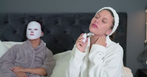 Multicultural friends enjoying home spa night using facial device and led sheet mask