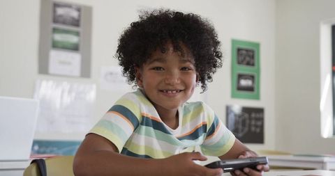 Smiling schoolboy holding handheld device at classroom desk, learning with technology