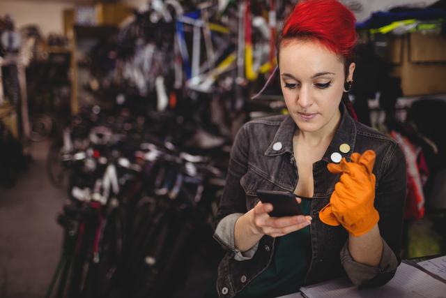 Female mechanic with red hair and denim jacket using mobile phone in a bicycle workshop. She is wearing orange gloves and surrounded by bicycles and tools. This image can be used for themes related to small businesses, women in trades, technology in the workplace, and professional mechanics.