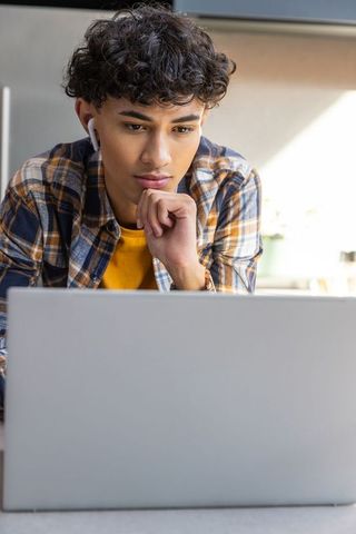 Young Man Concentrating on Laptop with Wireless Earbuds at Home