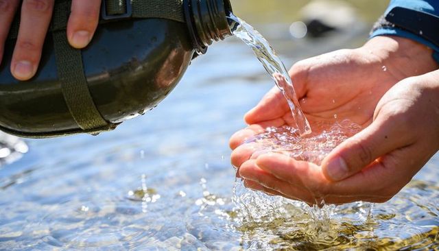 Pouring Water from Olive Green Canteen into Cupped Hands over Sunlit Stream