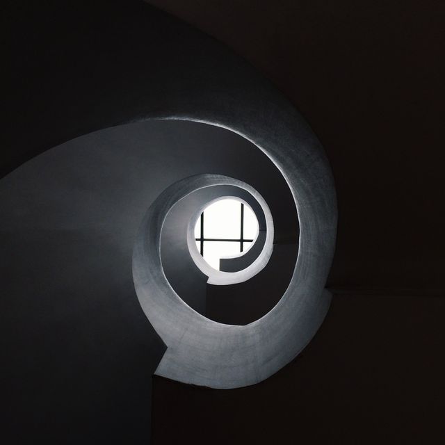 Elegant image of modern spiral staircase with natural light creating dramatic shadows and a minimalistic, abstract effect. Ideal for use in architectural presentations, interior design websites, minimalist art collections, creative backgrounds, and promotional materials showcasing contemporary aesthetics.