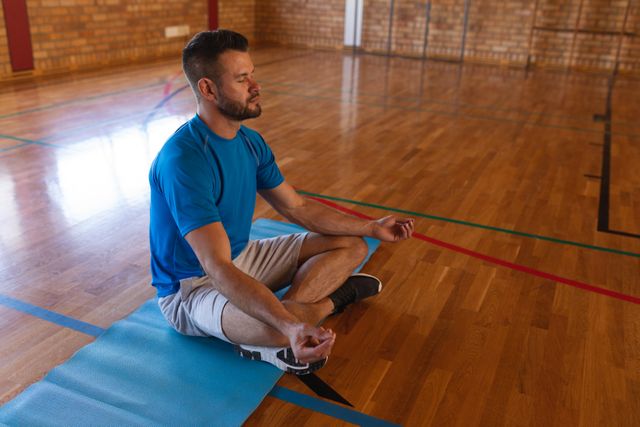Yoga teacher sitting cross-legged on a yoga mat in a school gym, meditating with eyes closed. Ideal for use in articles or advertisements about mindfulness, fitness, wellness, and healthy lifestyles. Can be used to promote yoga classes, meditation techniques, or school gym facilities.