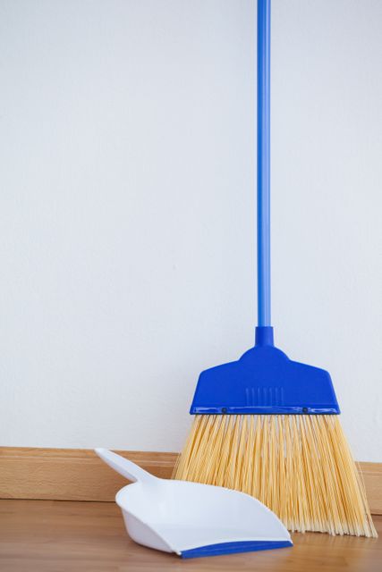 This image shows a close-up of a blue broom and a white dustpan placed on a wooden floor. It is ideal for use in articles, blogs, and advertisements related to housekeeping, cleaning products, home maintenance, and hygiene tips. The image can also be used in instructional materials on how to clean and maintain wooden floors.