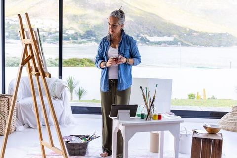 Senior artist studying canvas in waterfront home studio holding mug and palette