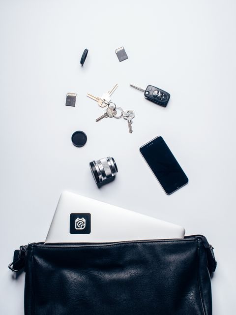This image illustrates a neat arrangement of everyday carry items such as a laptop, smartphone, keys, camera lens, SD card, and a black leather bag on a clean, white background. The items are typical essentials for professionals and creatives, making this image suitable for use in blog posts about productivity, work-life balance, and organization tips. It can also be used in advertisements for tech gadgets, minimalist backpacks, or professional gear accessories.