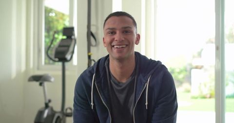Indian man smiling in home gym wearing navy hoodie near exercise bike bright interior