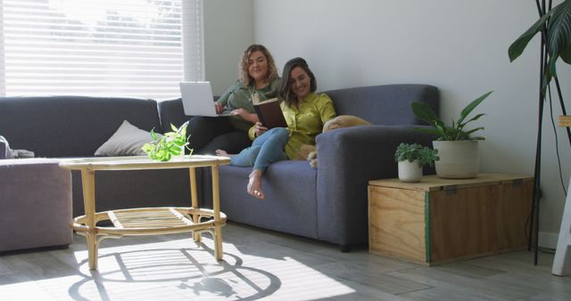 Two Women Relaxing on Couch Reading and Using Laptop in Bright Living Room - Download Free Stock Images Pikwizard.com