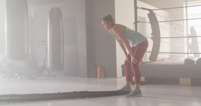 Woman taking break during high-intensity workout session in gym with boxing equipment and battle ropes. Suitable for themes related to fitness training, health, athleticism, and perseverance in personal workouts.