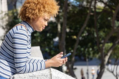 African american woman leaning on railing checking smartphone in sunny urban plaza