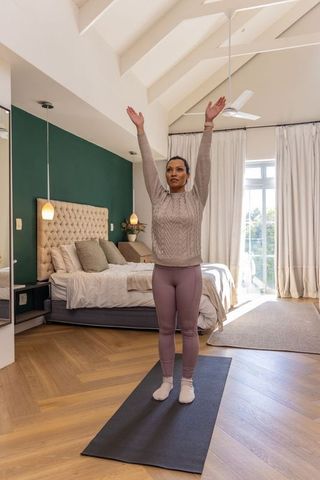 Woman stretching upward on yoga mat in sunlit modern bedroom with vaulted ceiling