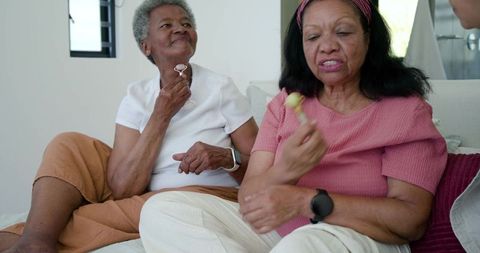Senior african american women relaxing on sofa using facial rollers and sharing candy