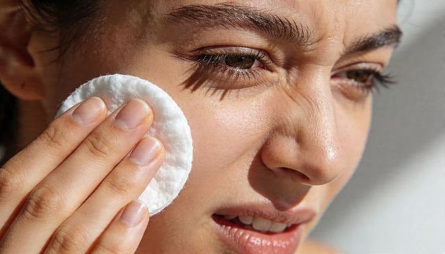 Closeup woman pressing cotton pad to cheek under natural window light skincare ritual