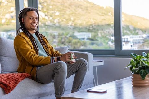 Relaxed african american man sipping coffee on modern sofa with scenic hillside window view