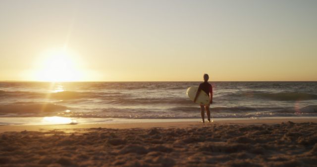 A lone surfer stands on a sandy beach and watches the sun set over the ocean. This image can be used for travel and tourism promotions, surf camp advertisements, summer vacation ideas, and relaxation themes. Ideal for website banners, social media posts, and marketing materials related to beach activities and coastal living.
