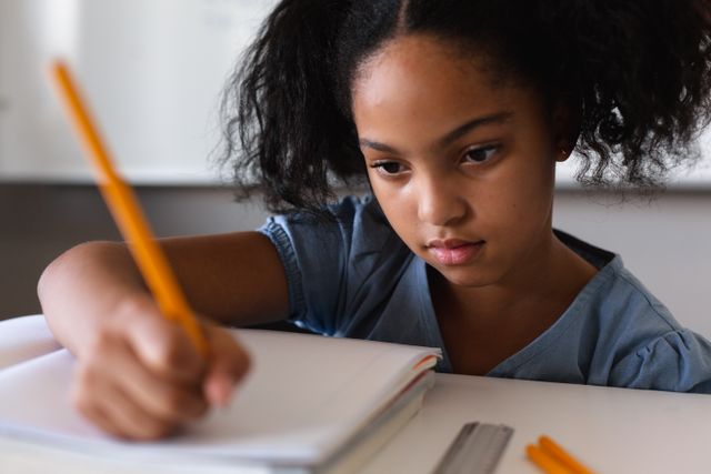 Biracial elementary schoolgirl writing on book while studying at desk in classroom. unaltered, education, learning, studying, concentration and school concept.