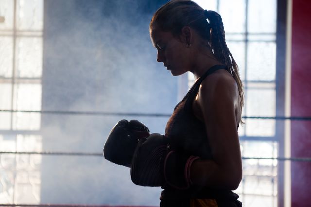 This image depicts a determined woman standing in a boxing ring in a fitness studio, wearing boxing gloves and looking focused. The scene is filled with a sense of strength and motivation, making it ideal for use in fitness and sports-related content, motivational posters, and advertisements for gyms or boxing classes.